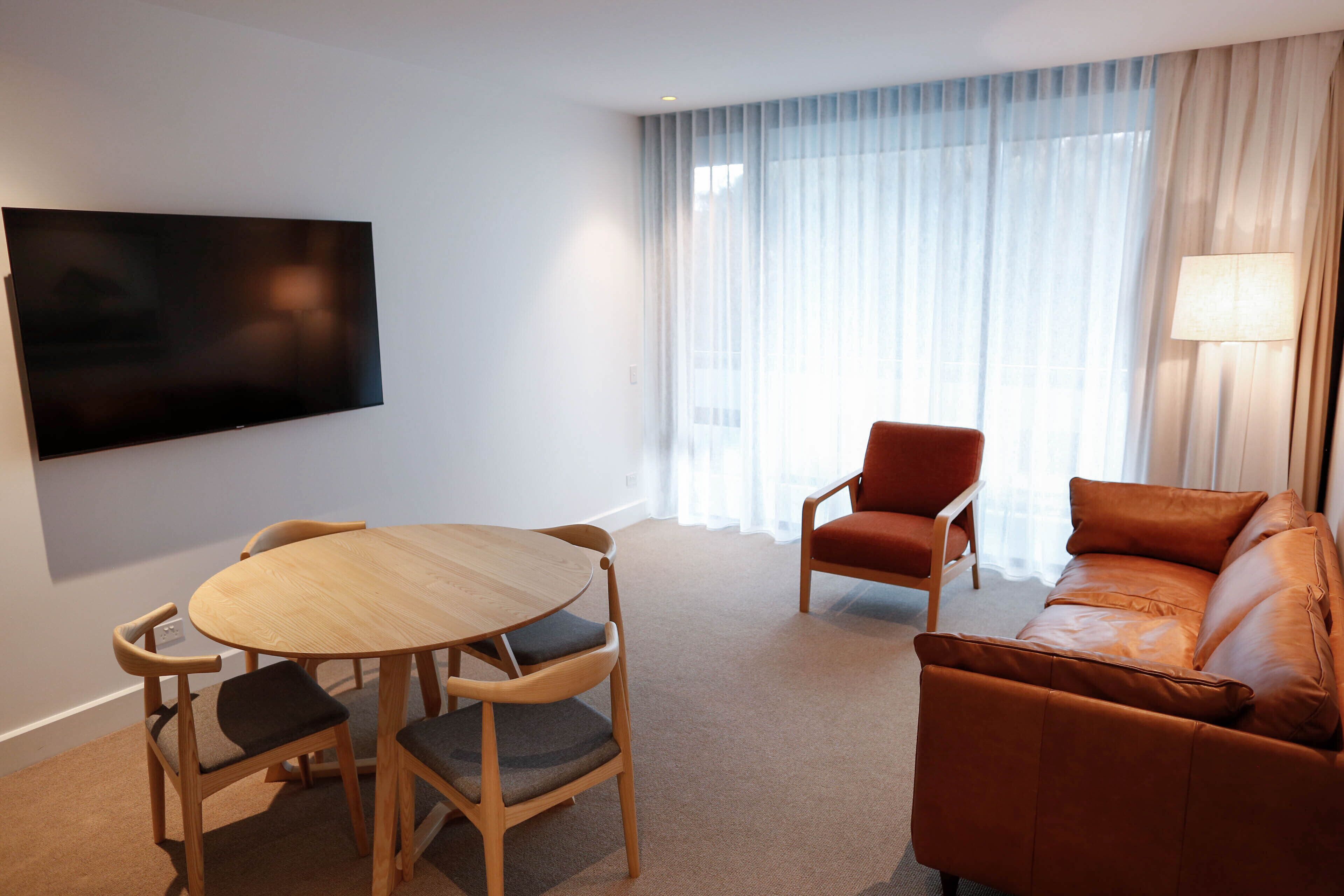 A modern living room with a round wooden table and four chairs, a leather sofa, a TV mounted on the wall, and a large window with sheer curtains.