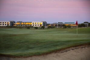 En golfbana med en röd flagga, moderna byggnader i bakgrunden och en klar himmel.