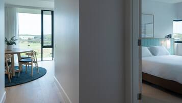 Habitación de hotel con cama, zona de comedor con mesa y sillas, y un amplio ventanal con vistas al exterior.