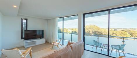 A modern living room with a large TV, comfortable seating, and a balcony with a view.