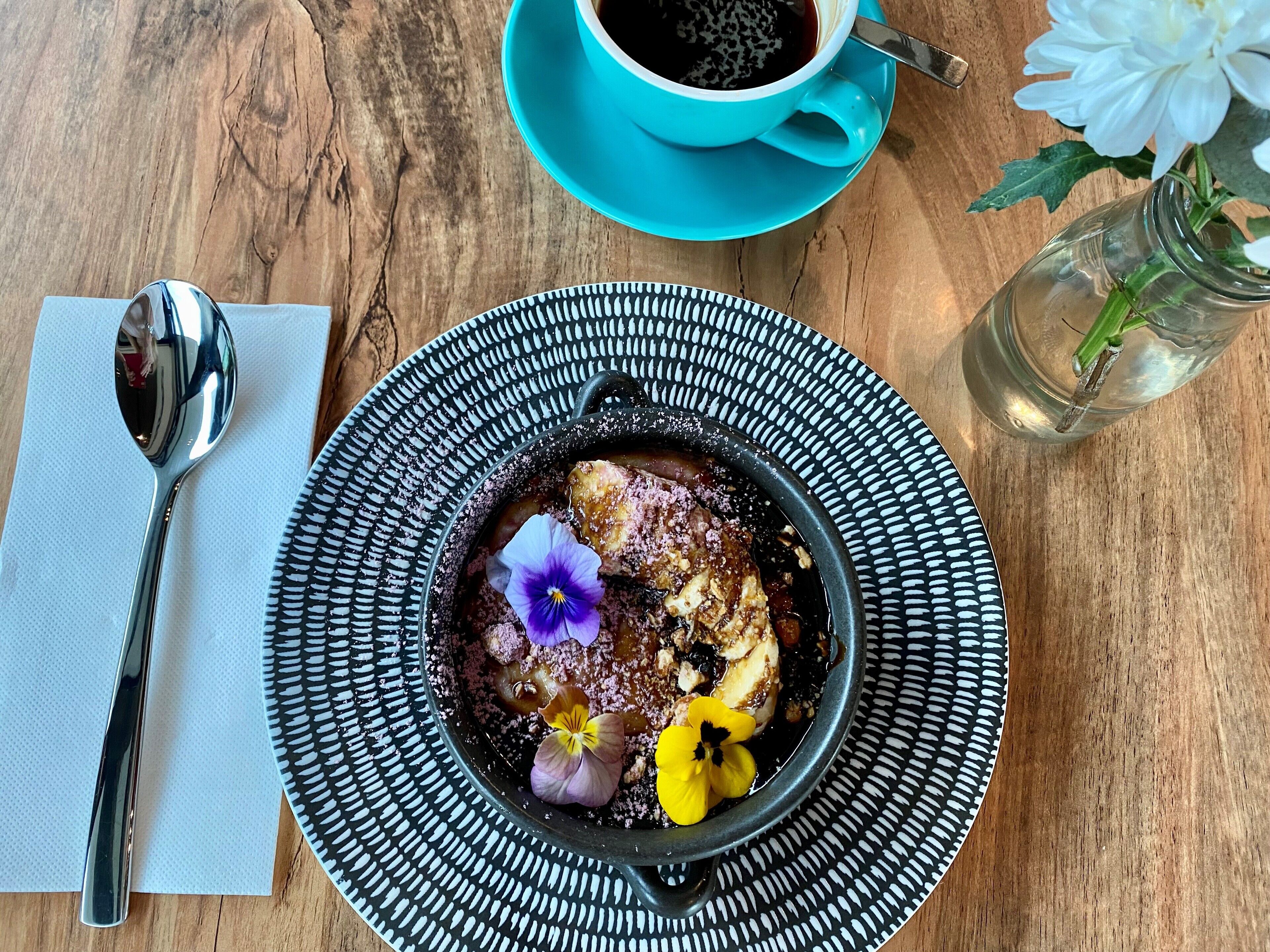 A dessert on a black plate with a spoon and a blue cup of coffee on a wooden table.