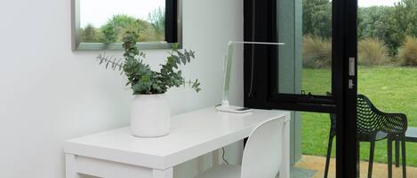 A white desk with a potted plant, a computer, and a chair in front of a window with a view of greenery.