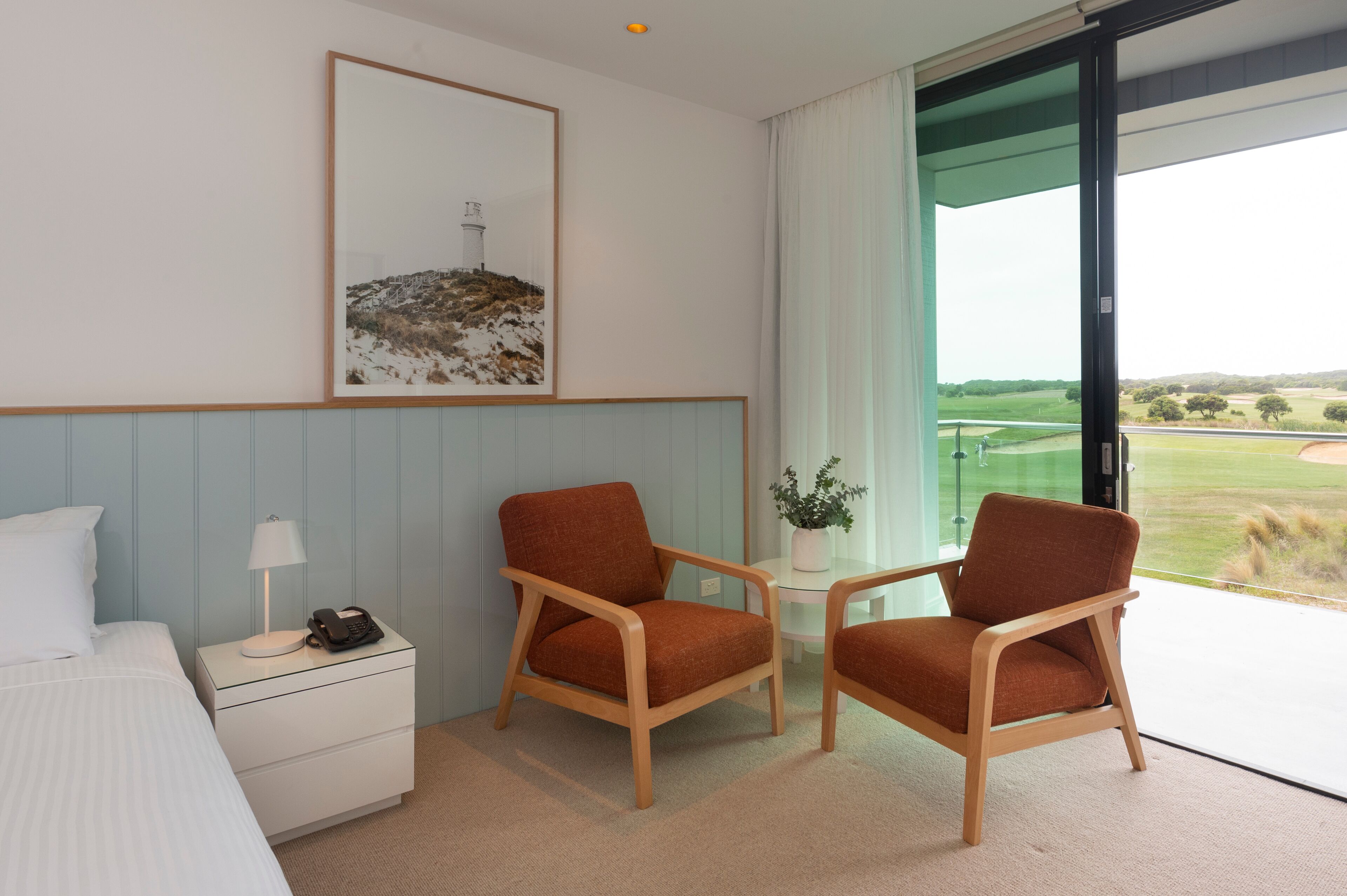 A hotel room with a bed, two armchairs, a nightstand with a lamp and phone, a framed picture on the wall, and a view of a golf course through a large window.