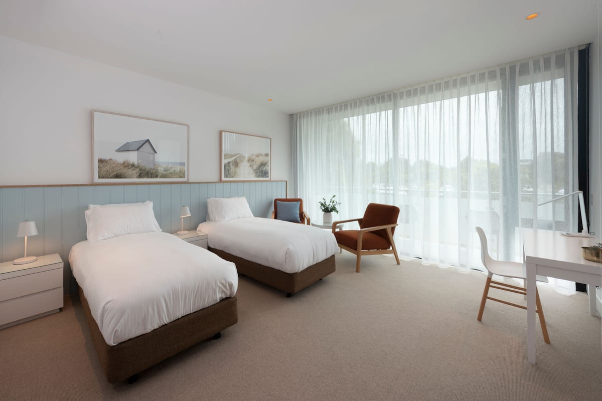 Ein Hotelzimmer mit zwei Betten, einem Schreibtisch, einem Stuhl und großen Fenstern mit transparenten Vorhängen.