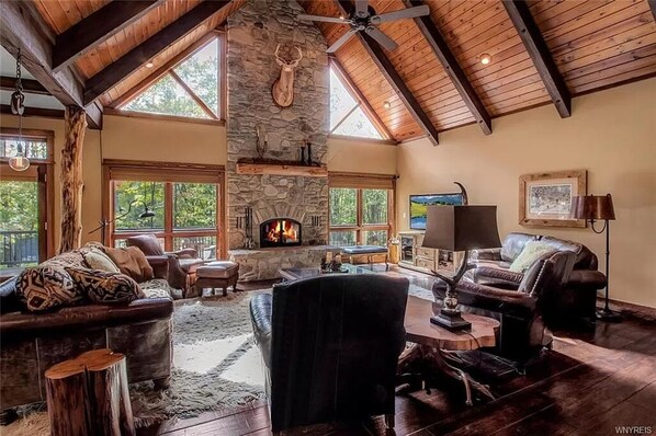 A spacious living room with a stone fireplace, leather furniture, and large windows offering a view of the outdoors. - Luxurious Chalet! Hot-tub, Bonfire & A+ Location for Skiing/Town (Ellicottville)