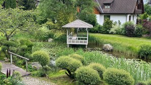 A serene garden with a wooden gazebo, a pond with lily pads, and houses in the background.
