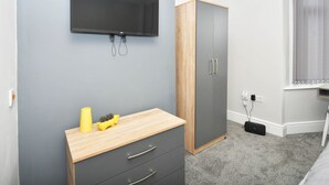 Une chambre avec un mur gris, un téléviseur fixé au mur, une table de chevet en bois avec des tiroirs, une armoire grise et une fenêtre avec des rideaux.