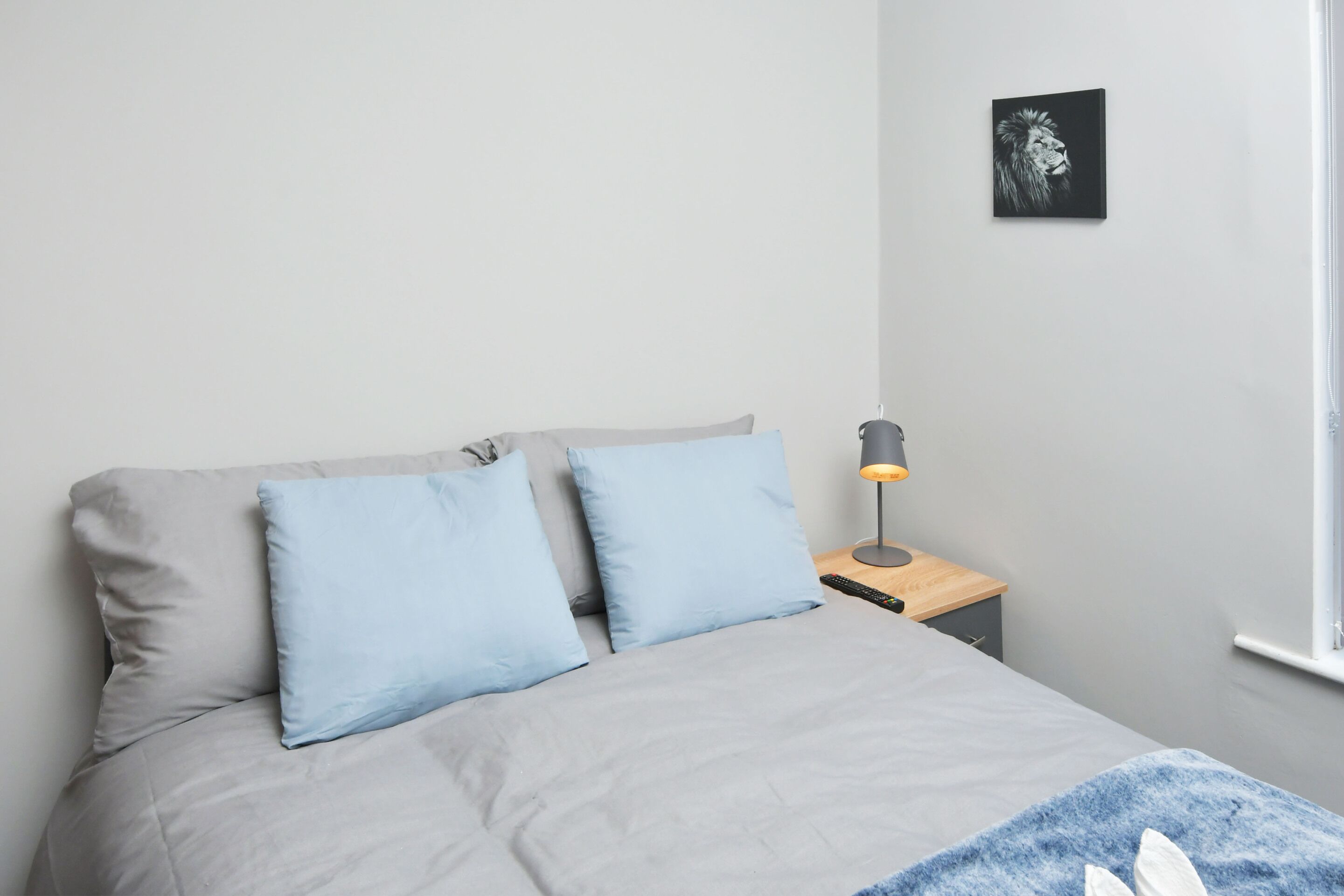 Una cama bien tendida con ropa de cama gris y azul, una lámpara de noche y un cuadro en la pared.