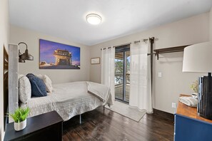 A bedroom with a bed, a nightstand, a lamp, a window with curtains, a painting on the wall, and a small plant. - Whiskey Hills At Terry Peak (North Lawrence)