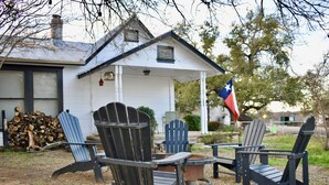 A white house with a porch, two Adirondack chairs, a fire pit, and a Texas flag. - “El Castellito” on the Llano River [4 Bed House] (Llano)