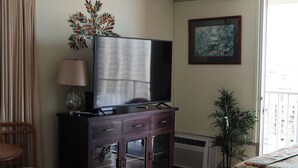 Un salón con un mueble de madera oscura, un televisor de pantalla plana, una lámpara, una silla, una planta en maceta y un cuadro en la pared.