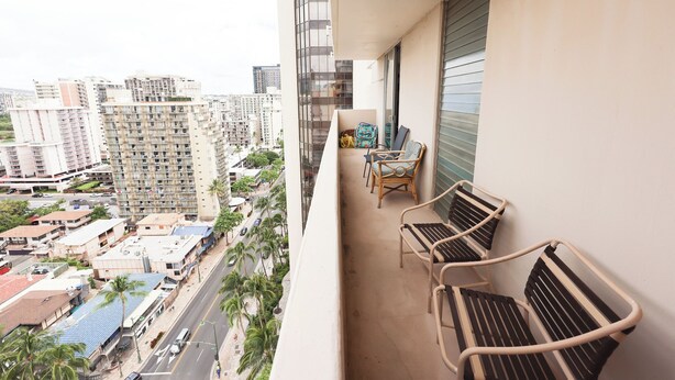 Un balcón con dos sillas de mimbre y una mesita pequeña, con vistas a un paisaje urbano de edificios altos y palmeras.