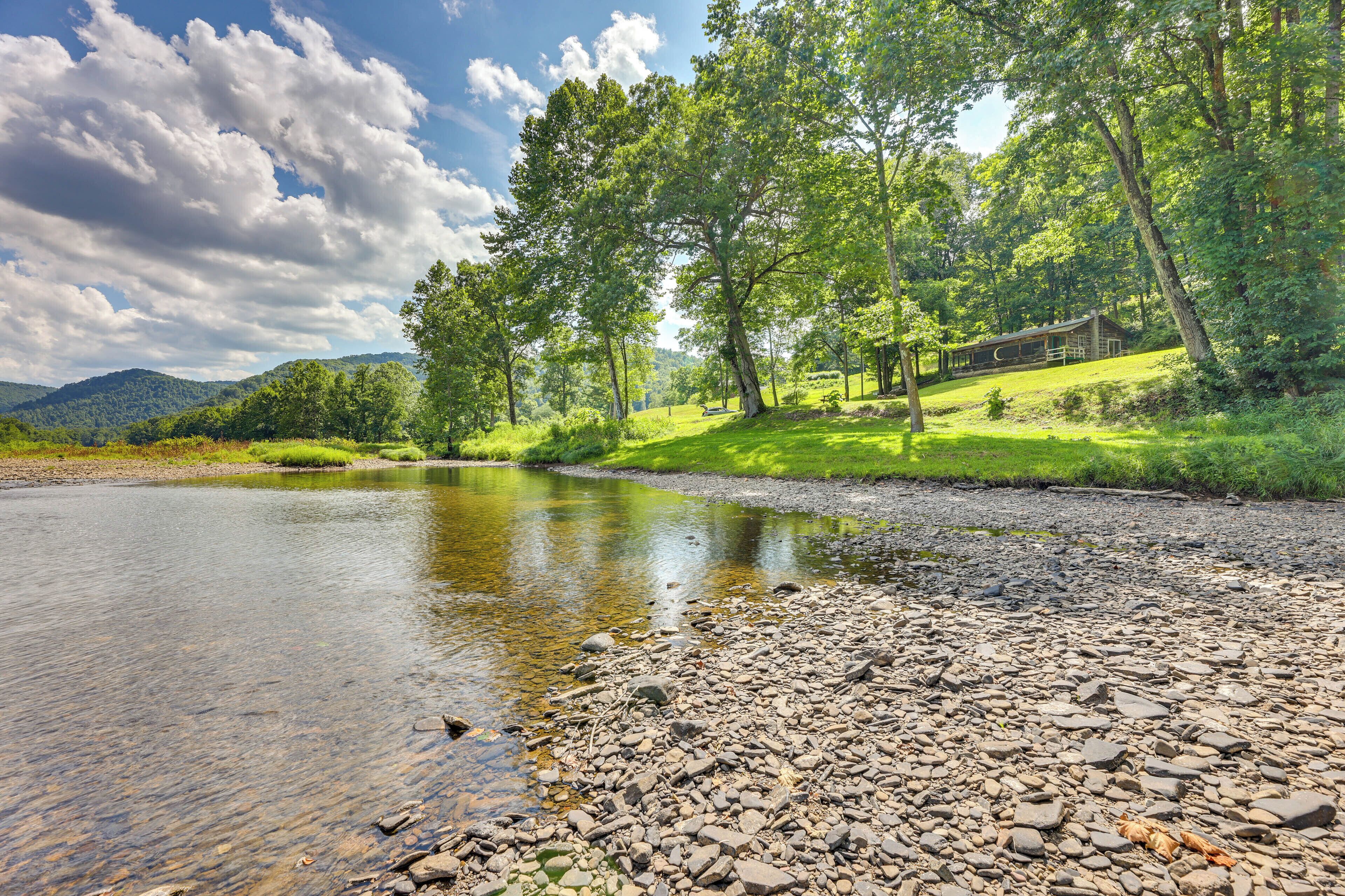 A riverside landscape with a wooden cabin, surrounded by trees and hills.