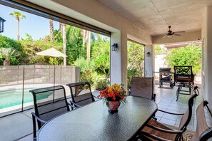 Un patio exterior con piscina, sillas de comedor, una mesa con un jarrón de flores y una parrilla.