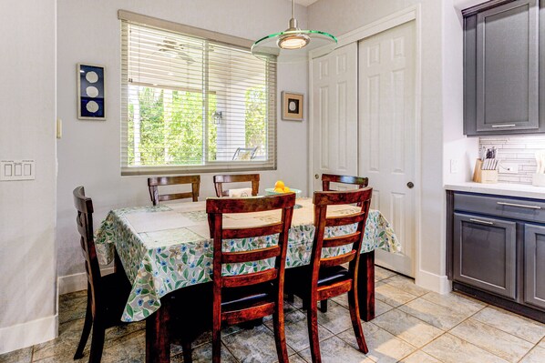 Un comedor con una mesa dispuesta para dos, sillas, una ventana con persianas y una zona de cocina con armarios.