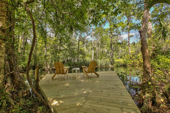 Una terraza de madera con dos sillas y una mesa pequeña, rodeada por un bosque frondoso y una masa de agua tranquila.