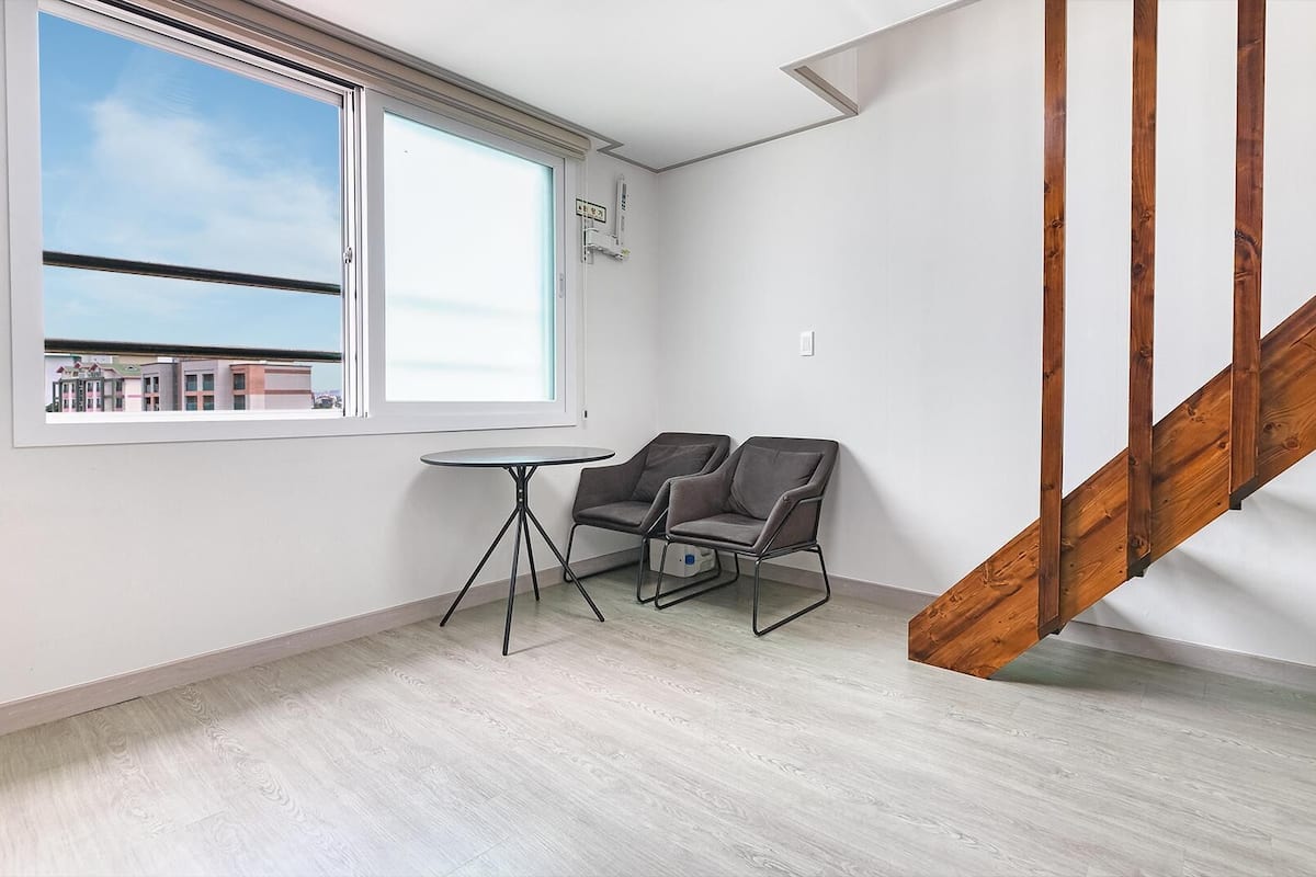 A room with a wooden staircase, a window with a city view, and two chairs near a small table.