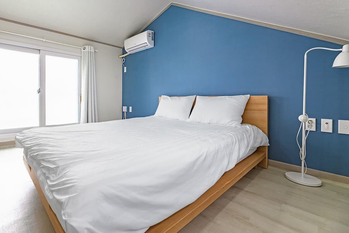 A bedroom with a bed, a window with curtains, a wall-mounted air conditioner, and a bedside lamp.