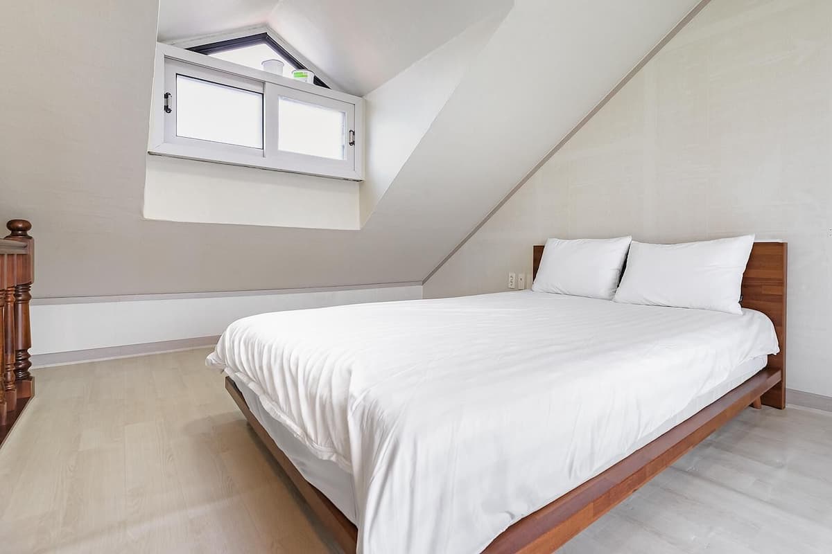 A bedroom with a sloped ceiling, a bed with white bedding, a wooden headboard, and a small window with a view of the sky.
