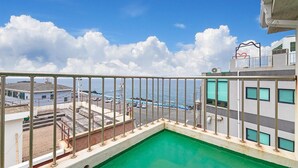 Un balcon donnant sur une piscine verdoyante, avec vue sur une zone côtière bordée de bâtiments et sur la mer.
