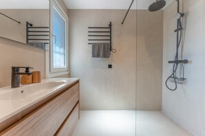 Une salle de bain moderne avec un grand miroir, un meuble-lavabo en bois, une douche équipée d’un pommeau de douche à main et un porte-serviettes.