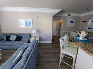 A living room with a grey sofa, a dining area with a white table and chairs, and a kitchen with a granite countertop.
