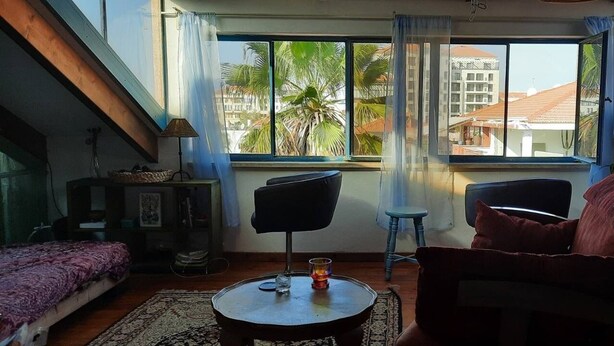 Una habitación con una cama, dos sillas, una mesita con un vaso y vistas a palmeras y edificios a través de amplios ventanales.