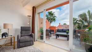 A modern living room with a sofa, a table, and a chair, a glass door leading to an outdoor patio with furniture and plants. - Apartment 'La Fragata Estepona 3b 2b' with Sea View, Wi-Fi and Air Conditioning (Estepona)