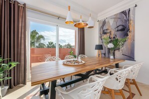 A dining area with a wooden table, white chairs, pendant lights, a painting, and a view of palm trees.