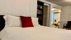 A neatly made bed with white and red pillows, a bookshelf with books, and a chair in a room with white walls and a door leading to another area. - A bright suite with lots of room for fun and relaxation, just 5 minutes from the Halifax ferry! (Dartmouth)