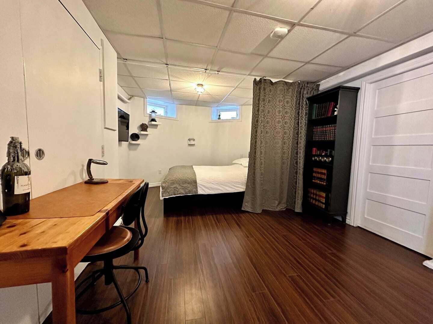 Ein kleines Schlafzimmer mit Holzschreibtisch, Bett, Bücherregal und Fenster mit Vorhängen.