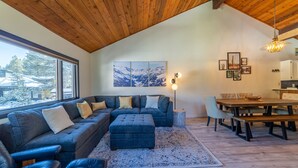 A modern living room with a sectional sofa, ottoman, dining table, and chairs. There is a large window with a view of snow-covered mountains. - Remodeled Meadowridge Family Retreat! Wifi, Spa (Mammoth Lakes)