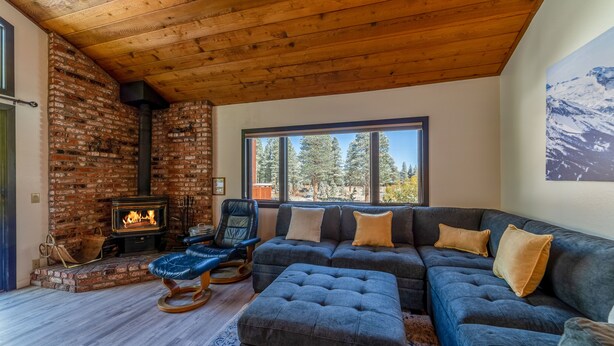 A cozy living room with a brick fireplace, a large window with a mountain view, a sectional sofa, and a wooden ceiling. - Remodeled Meadowridge Family Retreat! Wifi, Spa (Mammoth Lakes)