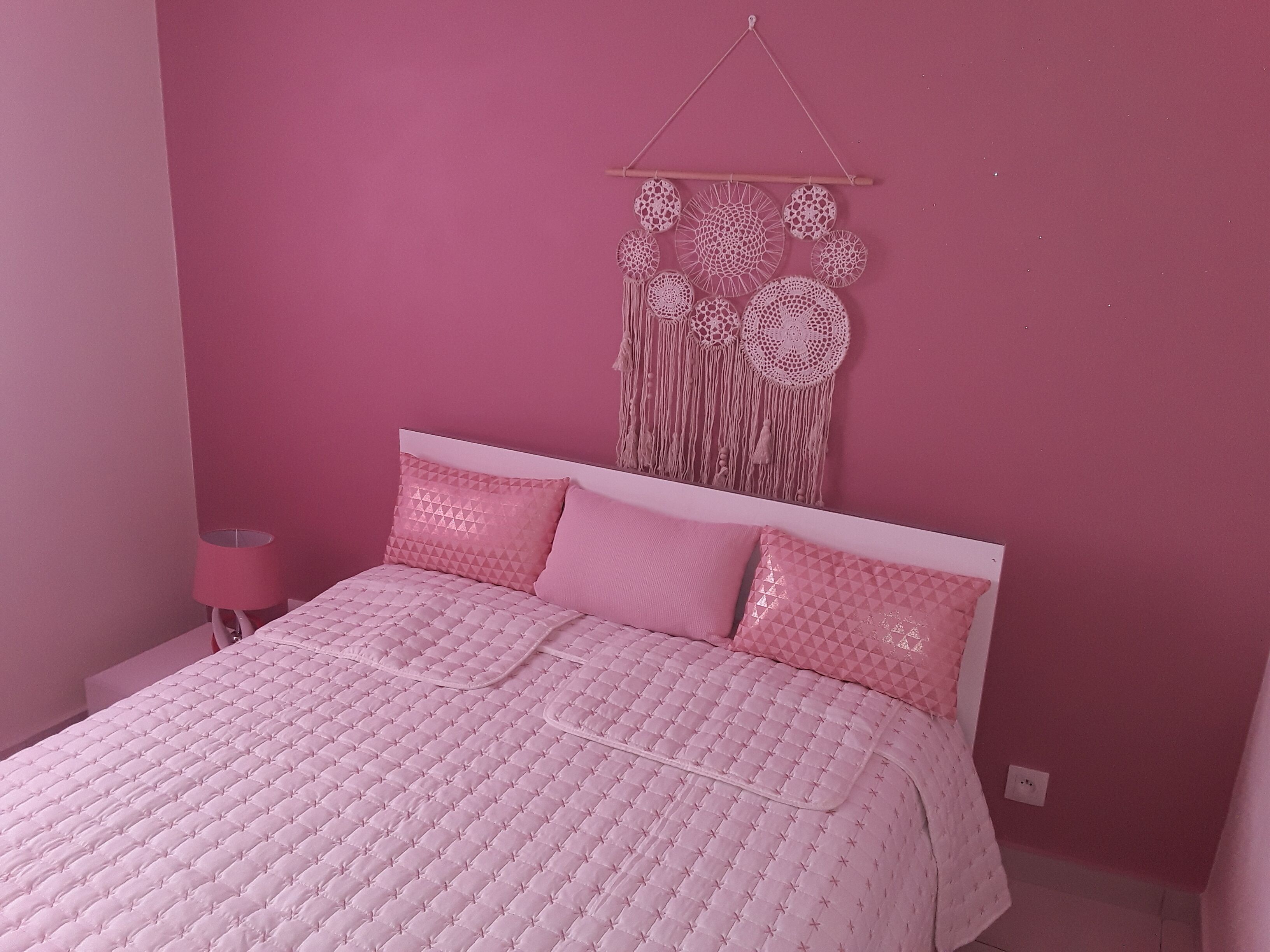 A pink-themed bedroom with a bed, pillows, a bedside lamp, and a wall hanging.