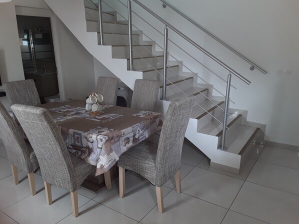 Une salle à manger avec une table dressée pour six personnes, entourée de six chaises. Un escalier avec une rampe en verre est visible à l’arrière-plan.