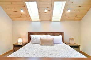A bedroom with a wooden ceiling, skylights, a bed with floral bedding, two bedside tables with lantern-style lamps, and a headboard.