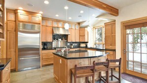 A spacious kitchen with wooden cabinets, a stainless steel refrigerator, a large island with bar stools, and a window providing a view of the outdoors. - Summit Rose Escape • Theater Rm • Gourmet Kitchen (Frisco)