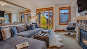 A modern living room with a grey sofa, a fireplace, and large windows offering a view of a house and trees. - Capitol Peak Lodge 3306 (Snowmass)