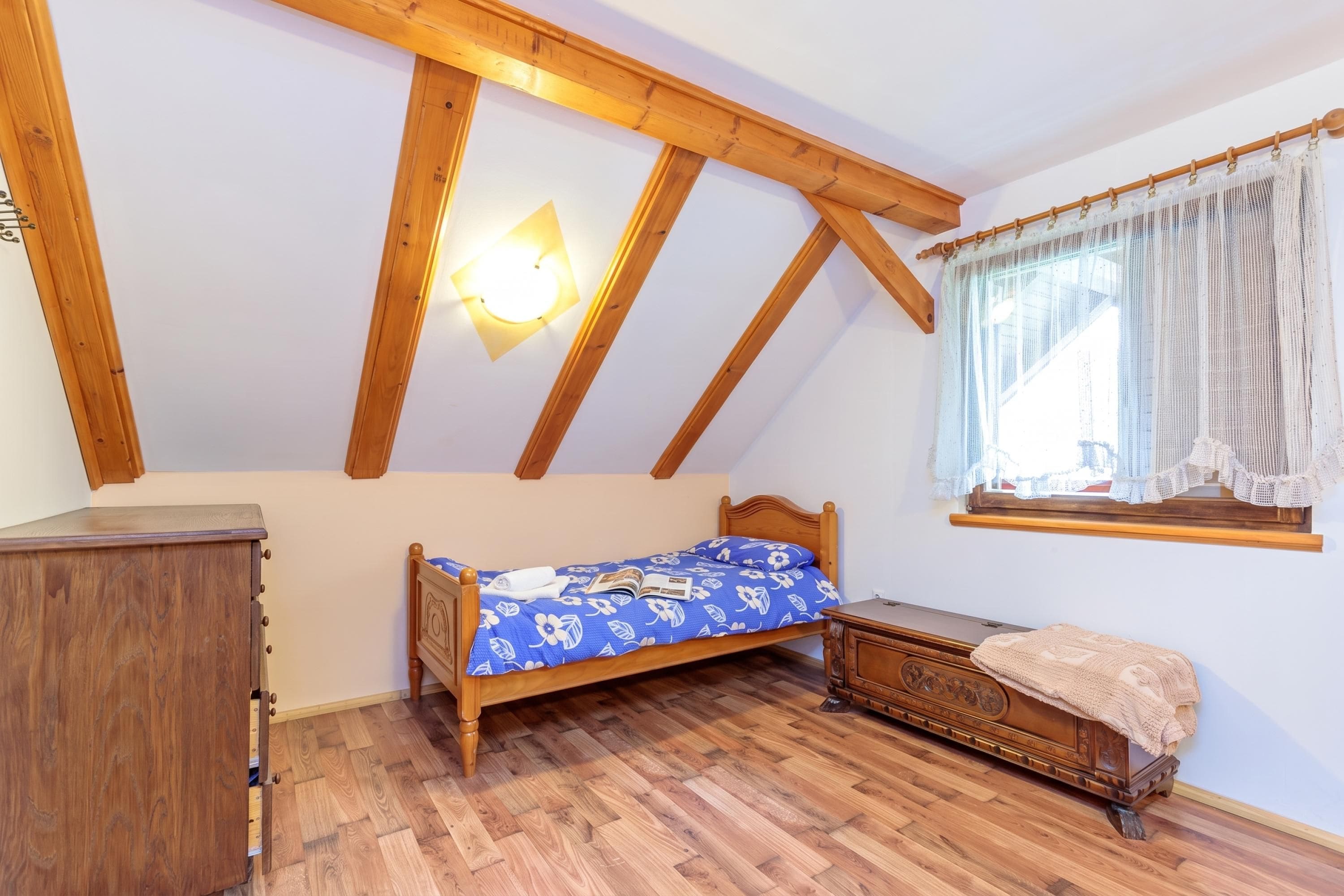 A bedroom with a wooden ceiling, a bed with a floral bedspread, a wooden dresser, and a window with sheer curtains.