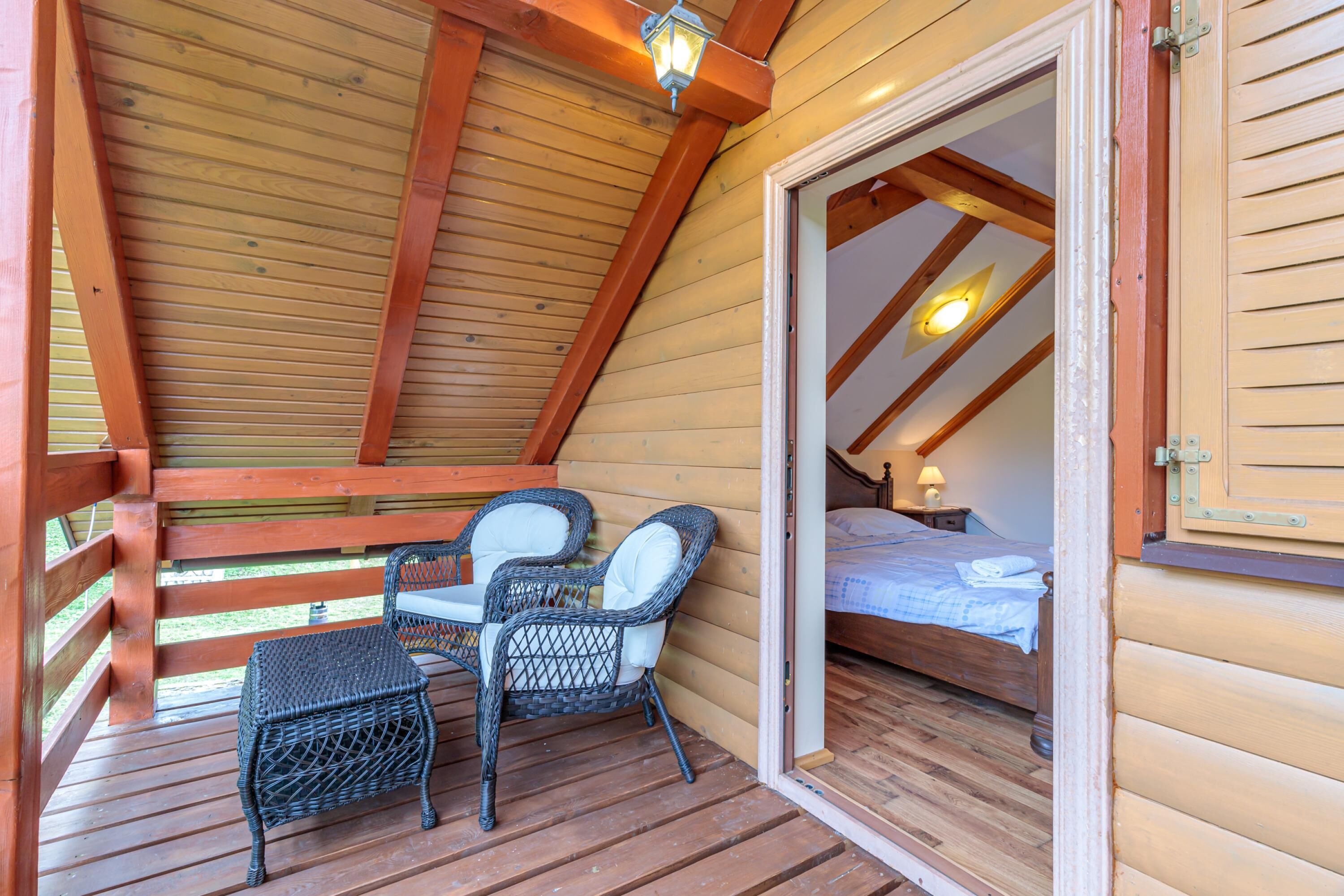 A wooden cabin with a balcony, wicker chairs, a bed, and a nightstand.