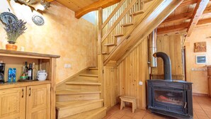 Een houten trap die naar een zolder leidt, een houtkachel, en een keuken met kasten en een plank.