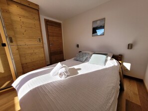 A neatly made bed with a folded towel, a wooden door, and a framed picture on the wall.