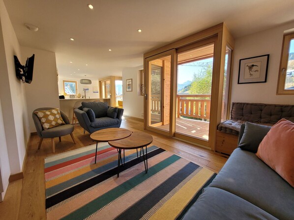A spacious living area with a sofa, armchairs, a coffee table, and a colorful striped rug. There is a sliding glass door leading to an outdoor balcony with a wooden railing.