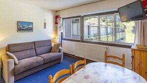 Quarto com sofá, mesa com toalha decorada com tema de café, televisão na parede e janela com vista para um edifício.