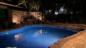 Une piscine avec un mur en pierre et un bord carrelé, entourée de plantes et bordée d’un chemin pavé.