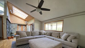 Un salon spacieux avec un grand canapé d’angle, un petit pouf et un ventilateur de plafond.