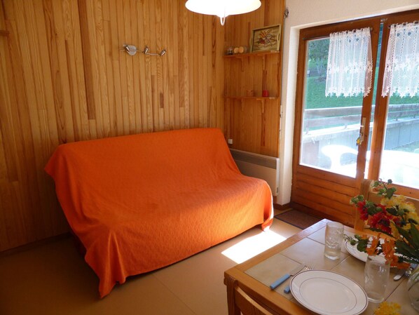 A room with a wooden wall, a covered sofa, a dining table with a vase of flowers, and a window with lace curtains. - 3-room apartment for 6 ppl, near slopes, parking (Combloux)