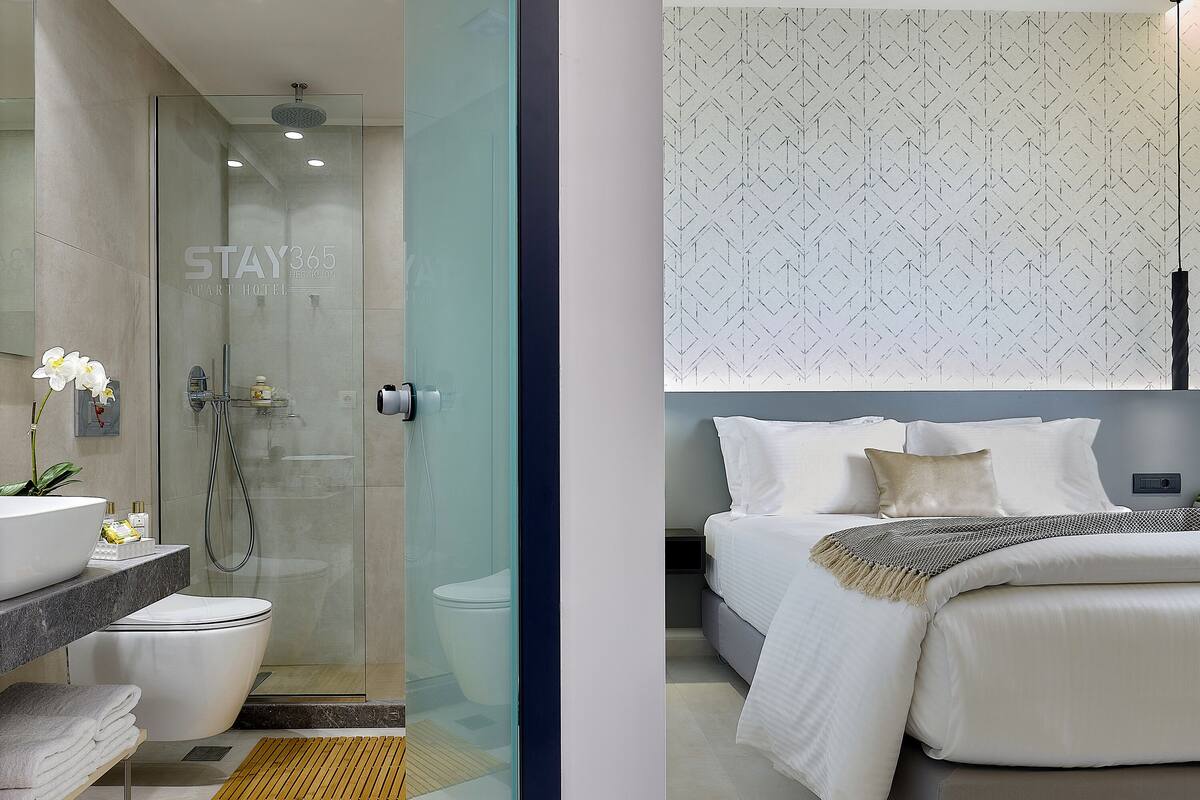 A modern bathroom with a glass shower enclosure, a white sink, and a toilet. The bedroom features a bed with white and beige bedding, a patterned wall, and a bedside table.