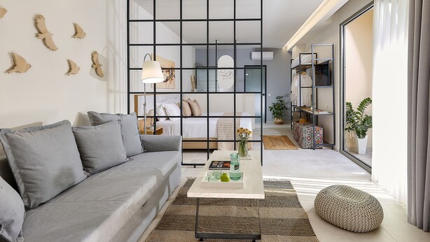 A modern living room with a grey sofa, a coffee table with decorative items, a black partition, and a view of a bedroom with a bed and a wardrobe. - Stay 365 Heraklion Apart Hotel (Heraklion)