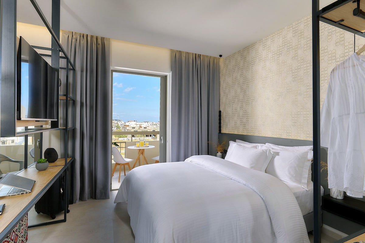 A modern hotel room with a large bed, a desk with a laptop, a small table with a plant, and a balcony with a view of the city.